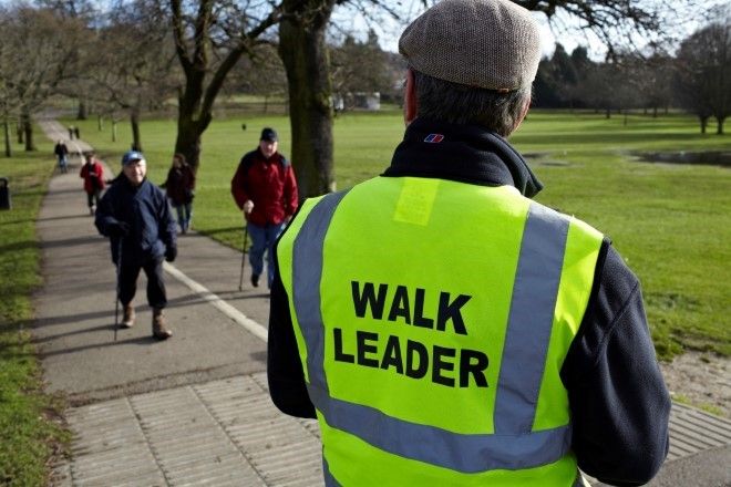 A walk leader in a hi-viz vest watches walkers walking past on a surfaced path in a park